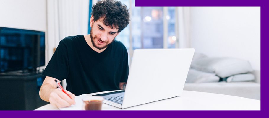 Imagem mostra um jovem de camiseta preta sentado atrás de uma mesa com um notebook a sua frente, fazendo anotações em uma folha.