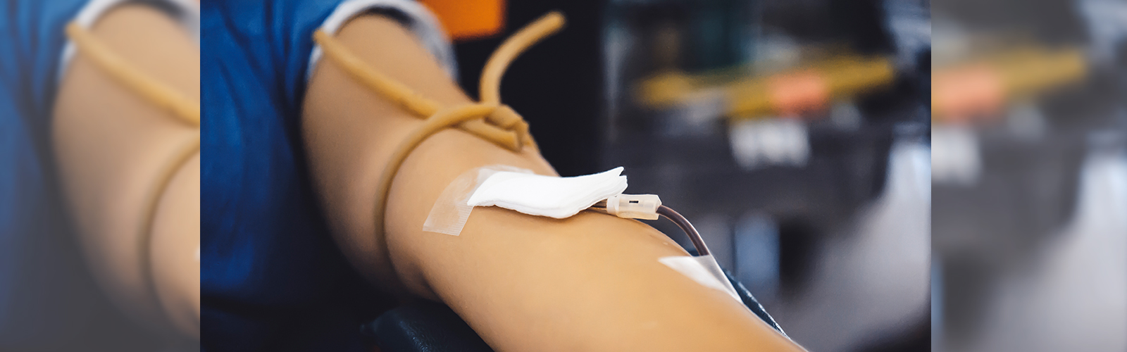 A imagem mostra um braço estendido durante o procedimento de doação de sangue, com uma agulha coberta por uma gaze inserida na pele.