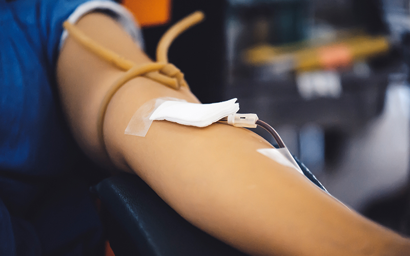 A imagem mostra um braço estendido durante o procedimento de doação de sangue, com uma agulha coberta por uma gaze inserida na pele.