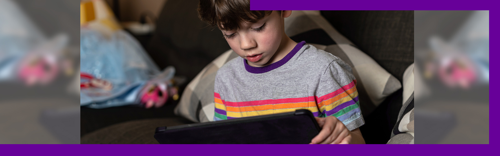 A imagem mostra um menino sentando no sofá, olhando para a tela de um tablet que está nas mãos dele.