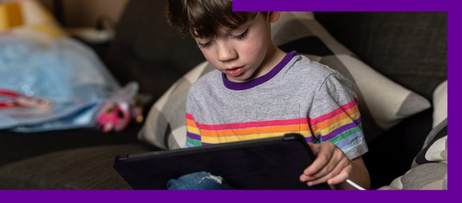 A imagem mostra um menino sentando no sofá, olhando para a tela de um tablet que está nas mãos dele.