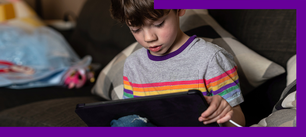 A imagem mostra um menino sentando no sofá, olhando para a tela de um tablet que está nas mãos dele.