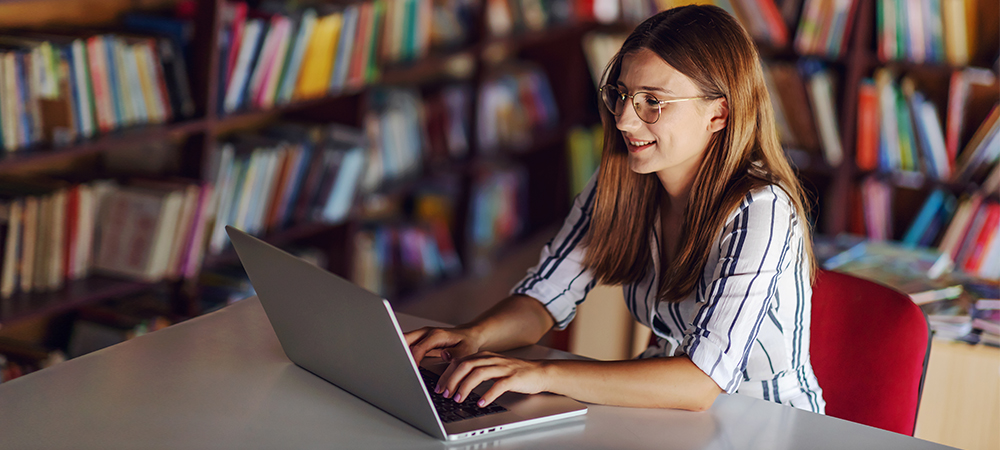 Para ilustrar pauta sobre curadoria de conteúdo, mulher jovem está sentada diante de um notebook com uma estante de livros de capas coloridas ao fundo. Ela tem cabelos loiros e lisos e usa óculos.
