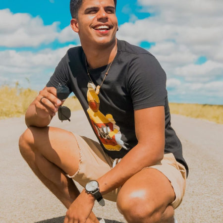 Elison Santos se destaca pelo ativismo indígena e aparece de camiseta e short, agachado em uma estrada com o céu azul em destaque.