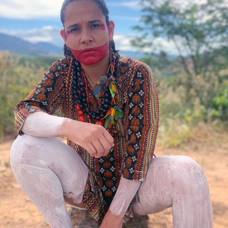 Shirley Djukurnã Krenak de destaca pelo ativismo indígena e aparece agachada, em meio a uma paisagem de cerrado, usando pintura tradicional, com parte do rosto pintado de vermelho e as pernas pintadas com tinta branca.