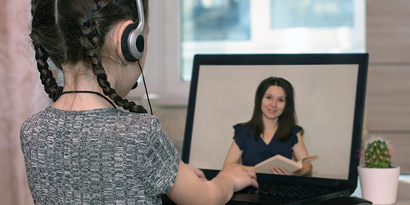 Menina usando tranças e com fone de ouvido está de costas para a foto e de frente para a tela de um notebook, onde uma professora faz a leitura de livros didáticos.