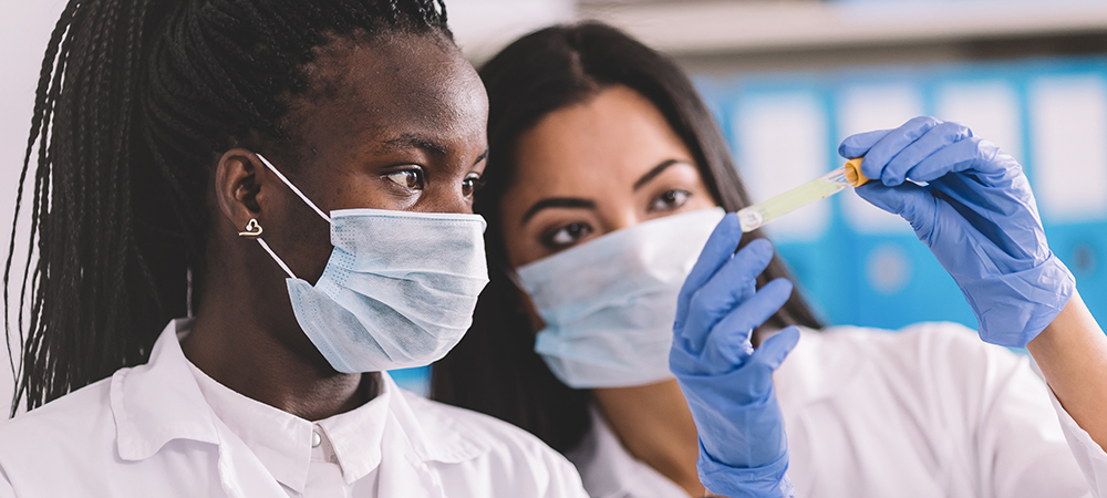A imagem mostra duas jovens, uma negra e uma branca em um laboratório. Ambas estão com jaleco branco e máscara de proteção e uma delas segura um tubo de ensaio.