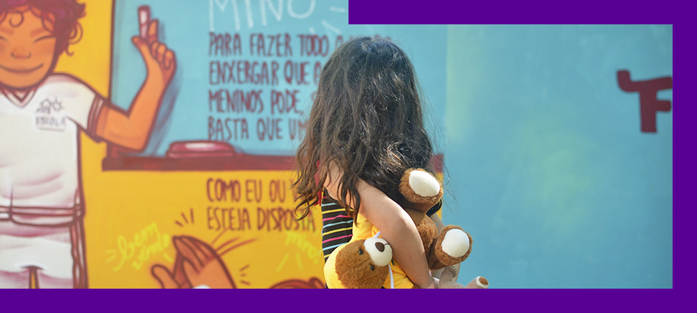 A imagem mostra uma criança de costas segurando um bicho de pelúcia olhando para o muro grafitado
