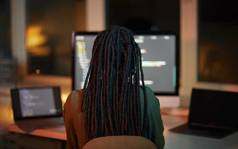 Mulher de dreads trabalha de costas. Na frente dela se vê um notebook e um computador.