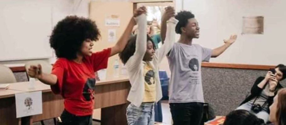 A imagem mostra três jovens do projeto Afroativos, que debate referências baseadas nos valores afro-brasileiros, de mãos dadas e erguidas em frente a estudantes em uma sala de aula.