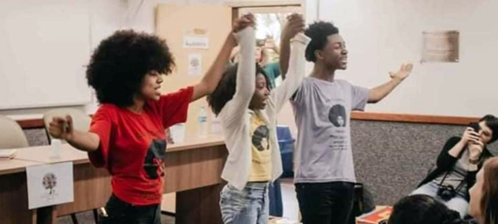 A imagem mostra três jovens do projeto Afroativos, que debate referências baseadas nos valores afro-brasileiros, de mãos dadas e erguidas em frente a estudantes em uma sala de aula.