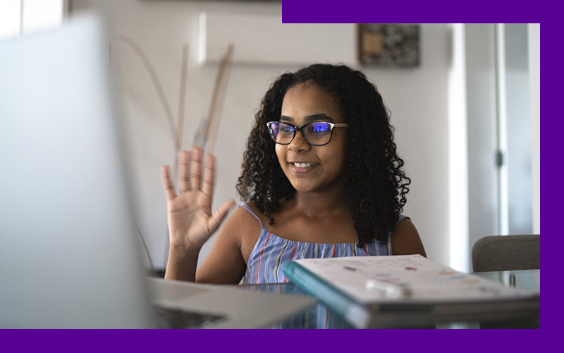 Imagem mostra uma menina sentada em frente a uma mesa onde está um notebook. Ela está sorrindo com uma das mãos levantadas.