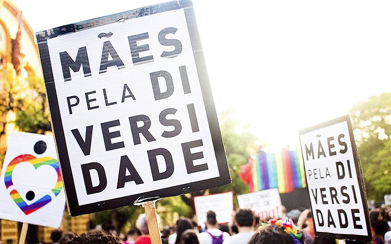 ONG Mães pela Diversidade atua no apoio a pais de filhos LGBTQIA+. Na imagem, cartaz traz o nome do coletivo em meio a uma passeata e a outros cartazes com as cores do arco íris.