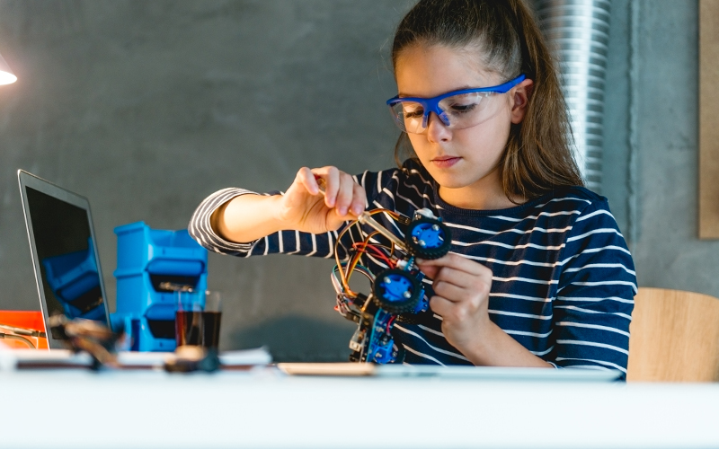 Menina está montando robô em uma bancada para representar pauta sobre a Coleção de Tecnologias Digitais. Ela usa óculos de proteção e tem os cabelos presos em rabo de cavalo.