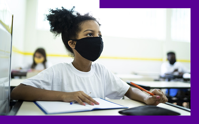 Imagem mostra uma menina dentro de uma sala de aula, sentada atrás de uma carteira escolar. Ela usa uma máscara de proteção preta, camiseta branca e segura um lápis na mão esquerda. Sobre a carteira há um caderno e um estojo.