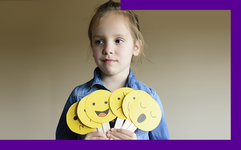 Em imagem que ilustra como ajudar as crianças a se expressarem, uma menina segura diversas placas que lembram emojis de expressões faciais, indicando sono, alegria, tristeza, entre outros. Ela está olhando para a frente com um sorriso discreto.
