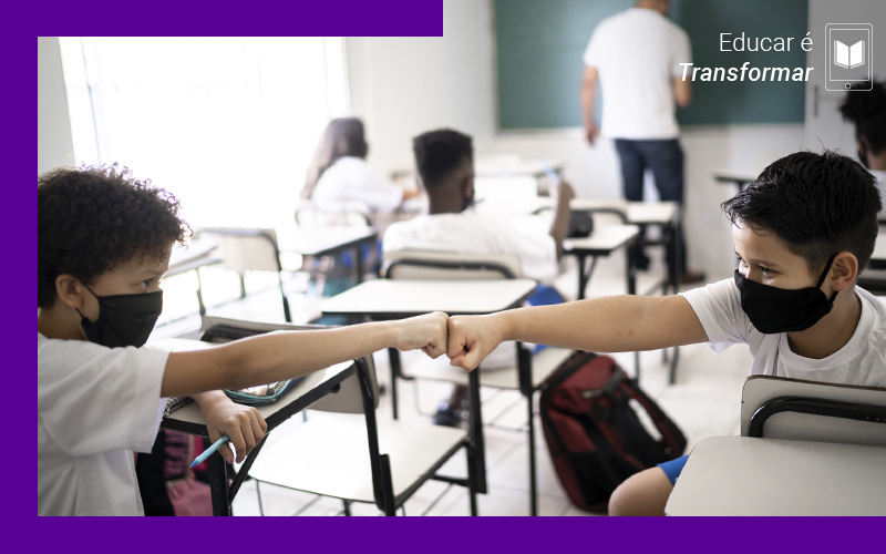 A imagem mostra duas crianças dentro de uma sala de aula, usando máscaras de proteção, se cumprimentando com as palmas das mãos fechadas.