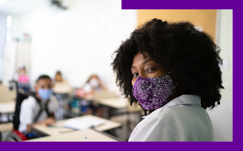 Imagem de uma professora negra em destaque dentro de uma sala de aula. Ela usa máscaras de proteção e os alunos sentados ao fundo também.