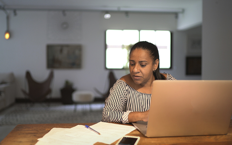 Mulher na sala de uma casa, sentada em frente a um computador, olhando para uns papeis sobre a mesa.