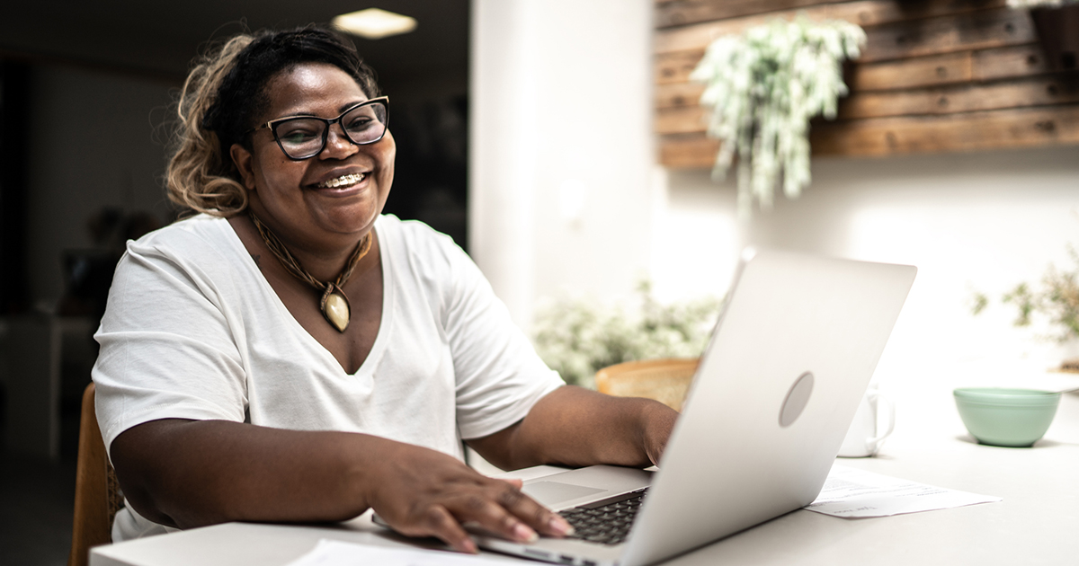 Imagem mostra uma mulher negra, de camiseta branca e um colar. Ela está sentada em frente um computador, sorrindo enquanto olha para a tela.