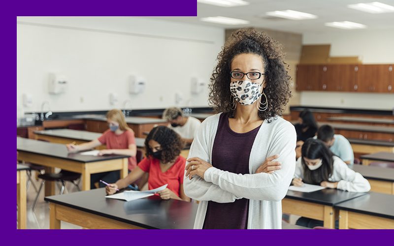 Imagem de uma professora de pé, com os braços cruzados, em uma sala de aula com alunos ao fundo, sentados em carteiras escolares. Todos estão usando máscara de proteção.