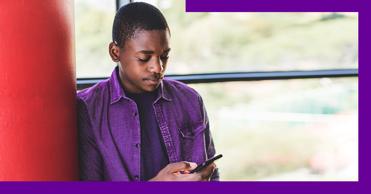 Imagem de um jovem negro, de camisa púrpura, encostado na parede, olhando para a tela de um celular que está nas mãos dele