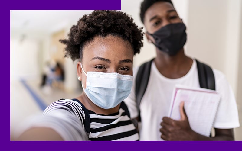 Imagem mostra em destaque dois estudantes jovens no corredor da escola, usando máscara de proteção individual