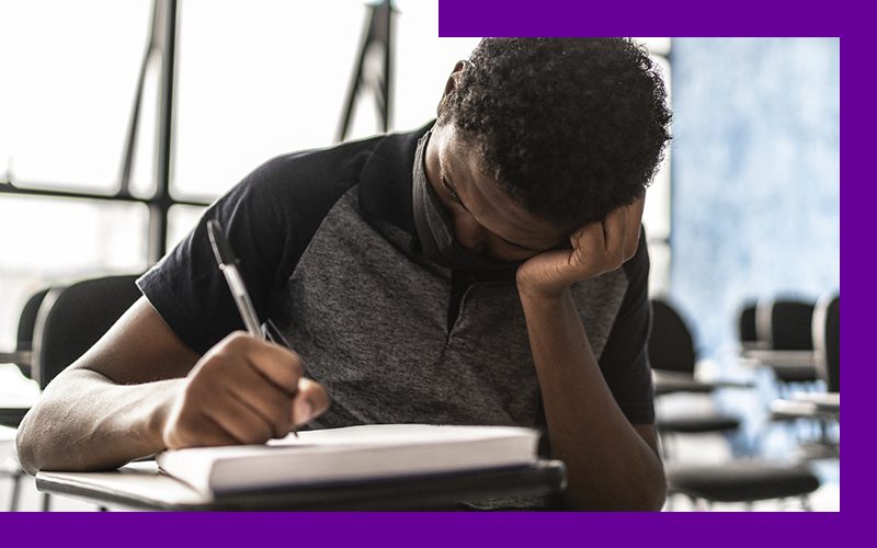 imagem de um jovem estudante na sala de aula, sentando em uma carteira escolar. Ele usa uma máscara de proteção individual e está com uma das mãos apoiada na cabeça, enquanto escreve em um caderno.