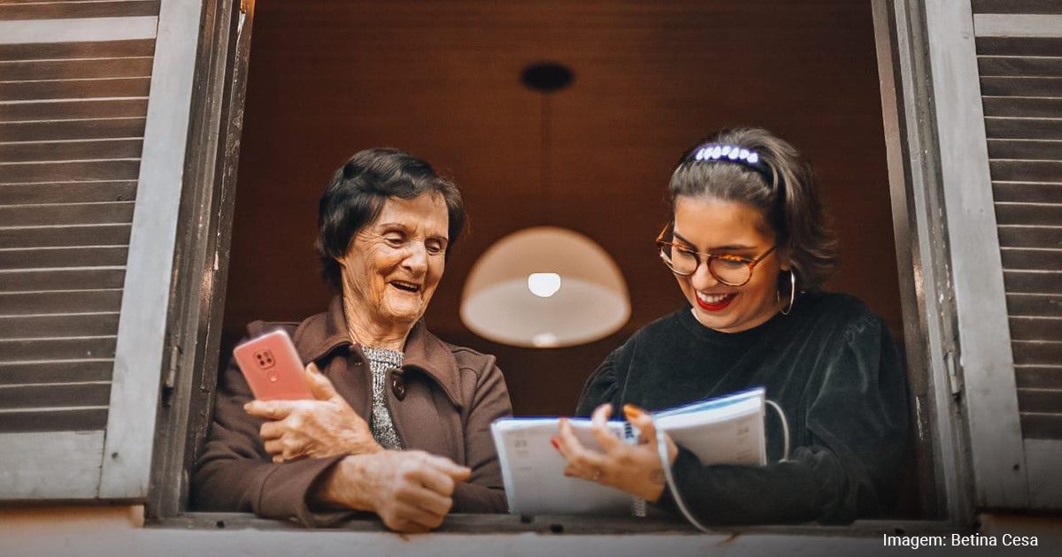 Imagem com Barbara Correa, que segura um caderno, e a sua avó Grete, com um celular nas mãos, na janela de casa