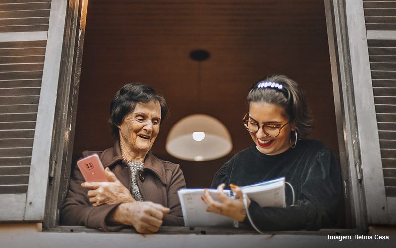 Imagem com Barbara Correa, que segura um caderno, e a sua avó Grete, com um celular nas mãos, na janela de casa