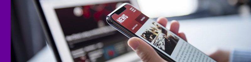 Imagem de uma pessoa segurando o celular ao lado de um computador com um site de notícias aberto