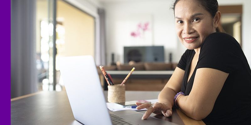 Imagem de uma mulher olhando para a câmera ao lado de um notebook na mesa