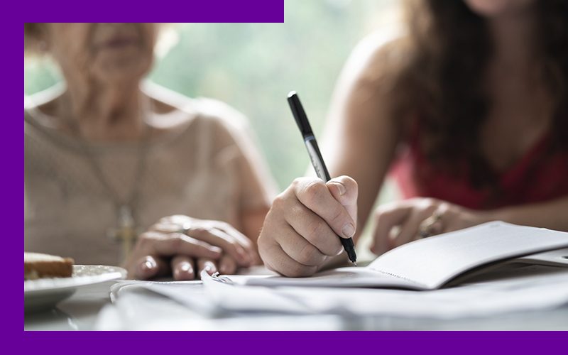imagem de duas mulheres lado a lado, uma senhora e uma jovem, com destaque para as mãoes da jovem escrevendo em um caderno.