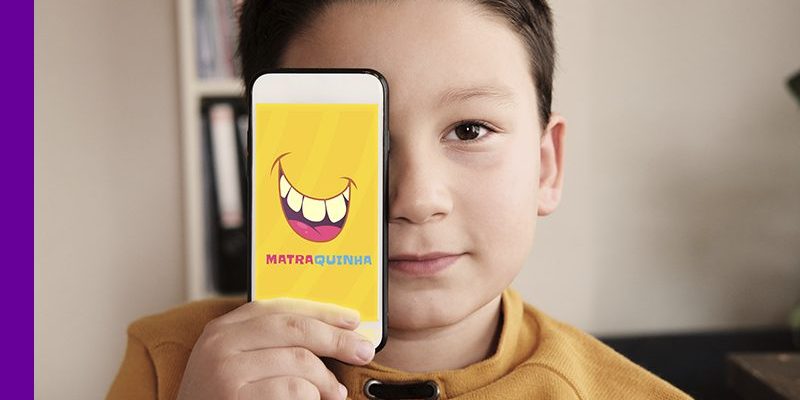 imagem mostra uma criança com um celular na mão, onde se vê o nome do aplicativo Matraquinha