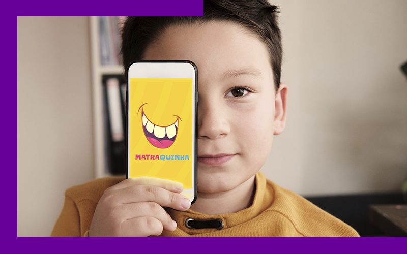 imagem mostra uma criança com um celular na mão, onde se vê o nome do aplicativo Matraquinha