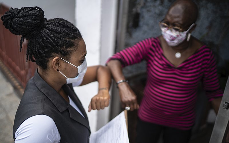 Imagem mostra duas mulheres de máscaras de proteção tocando os cotovelos para se cumprimentar durante a pandemia