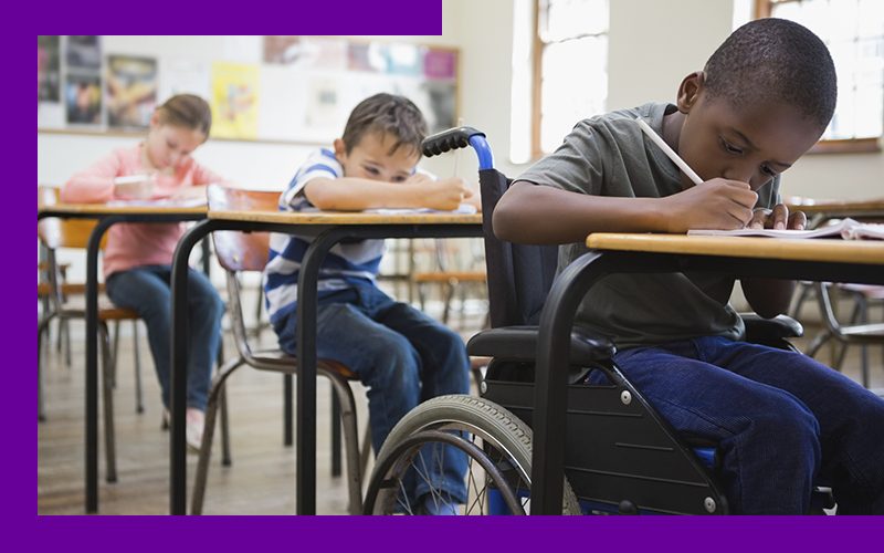 Imagem mostra três crianças estudando em sala de aula. Uma das crianças é negra e está em uma cadeira de rodas