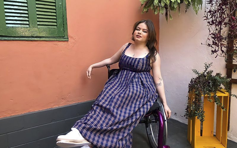 Imagem mostra a jovem fotógrafa e modelo usando um vestido xadrez, sentada em uma cadeira de rodas em um local com plantas