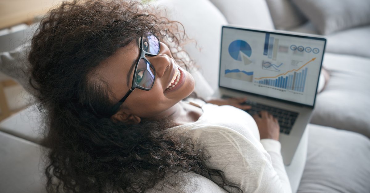 Imagem mostra uma jovem usando um computador. Ela é negra, tem cabelos compridos, usa óculos, está sentada em um sofá e está sorrindo.