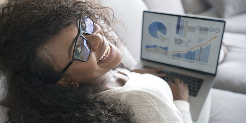 Imagem mostra uma jovem usando um computador. Ela é negra, tem cabelos compridos, usa óculos, está sentada em um sofá e está sorrindo.