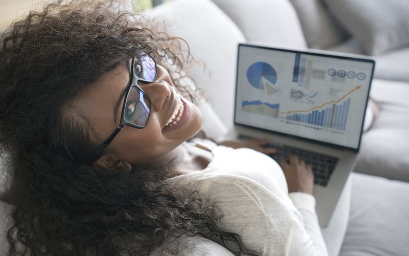 Imagem mostra uma jovem usando um computador. Ela é negra, tem cabelos compridos, usa óculos, está sentada em um sofá e está sorrindo.