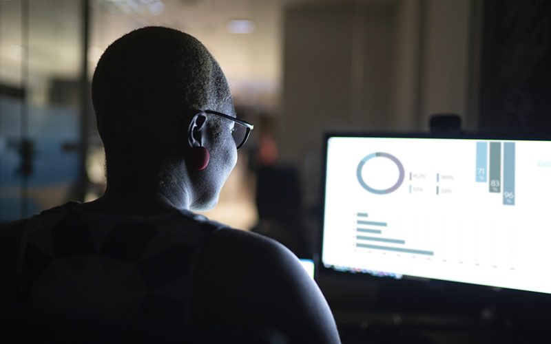 Imagem mostra uma jovem negra, ela usa óculos e está olhando para a tela do computador. A imagem capta a cena por trás da moça.