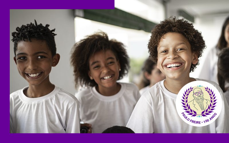 Imagem mostra três crianças sorrindo. No canto inferior direito da imagem há o selo comemorativo do centenário de Paulo Freire