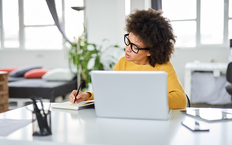 Imagem mostra mulher de óculos estudando em frente a um notebook e usando um caderno e caneta