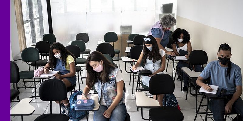 Imagem mostra estudantes em sala de aula sendo orientados por uma professora