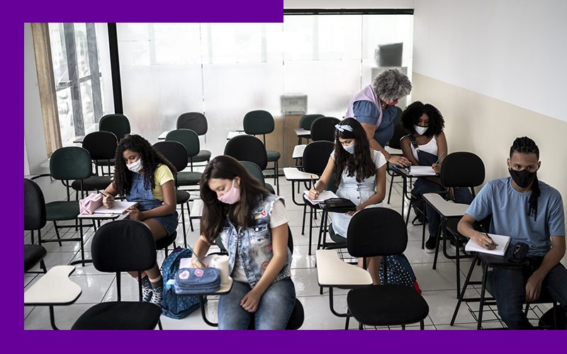 Imagem mostra estudantes em sala de aula sendo orientados por uma professora