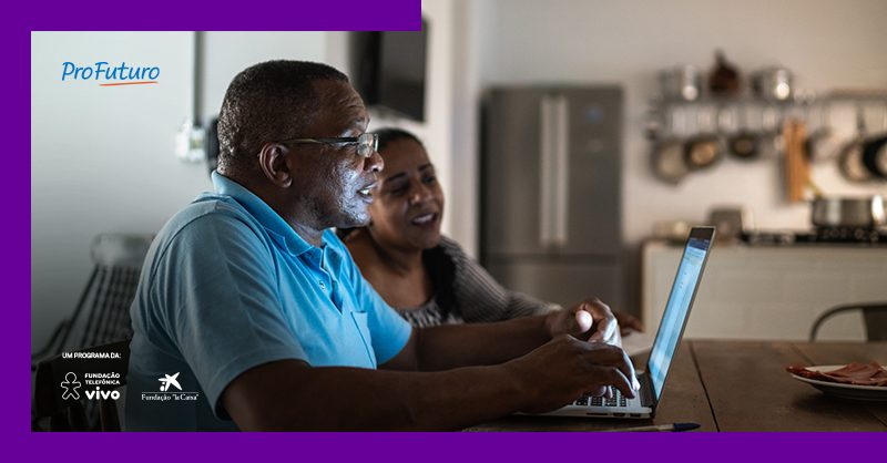 Imagem mostra um homem e uma mulher negros utilizando um computador. Eles estão em casa. O homem veste uma camisa azul. A mulher usa uma blusa cinza e tem cabelos compridos.