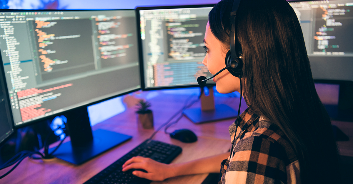 Imagem mostra uma jovem em frente a dois computadores. Ela está digitando e na tela há linguagens de programação.