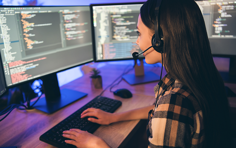 Imagem mostra uma jovem em frente a dois computadores. Ela está digitando e na tela há linguagens de programação.