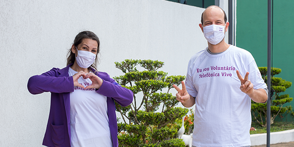 Imagem mostra um homem e uma mulher. Eles usam máscara e a camiseta do dia dos voluntários. Eles sorriem para a foto e fazem sinais com as mãos.